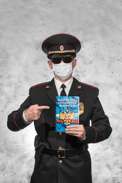 Russian police officer in uniform in a white medical mask and shows the index finger on the Constitution in the left hand on which is written in russian 