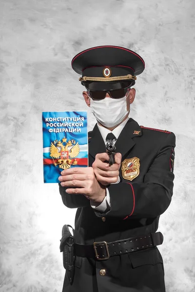 Russian police officer in uniform in a white medical mask and black glasses holds a gun in the right hand and the Constitution on which is written in russian \