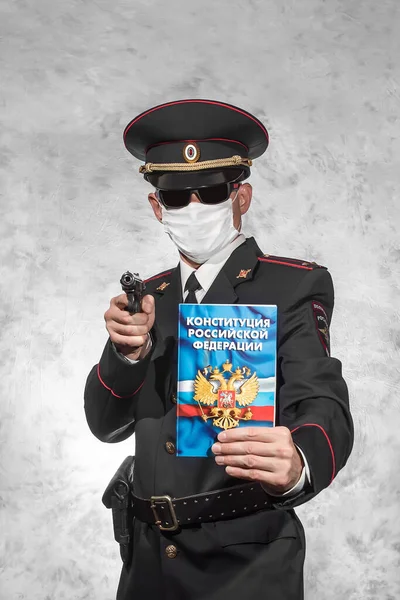 Russian police officer in uniform in a white medical mask and black glasses holds a gun in the right hand and the Constitution on which is written in russian \