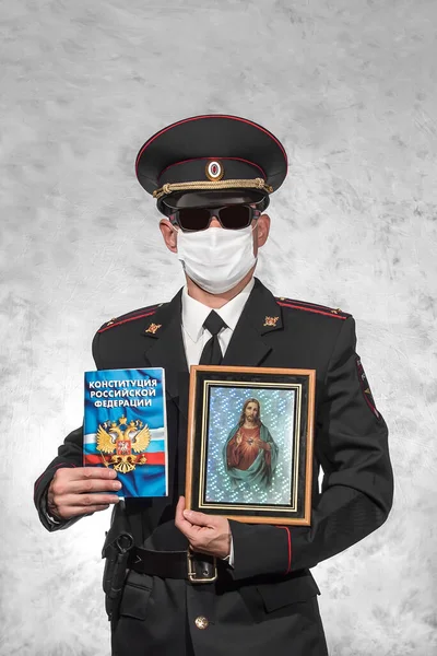 Russian police officer in uniform in a white medical mask and black glasses and holds Jesus Christ icon and the Constitution on which is written in russian 