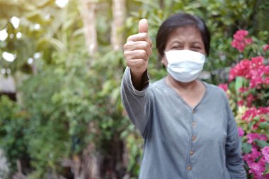 Tayland 'da Covid-19 veya Corona virüsünü önlemek için beyaz bez maskeli yaşlı bir Asyalı kadın ve Hava Kirliliğinin korunması için başparmaklar yukarı. Yaşlıların sağlığı konsepti. Kapalı ellere odaklanma.