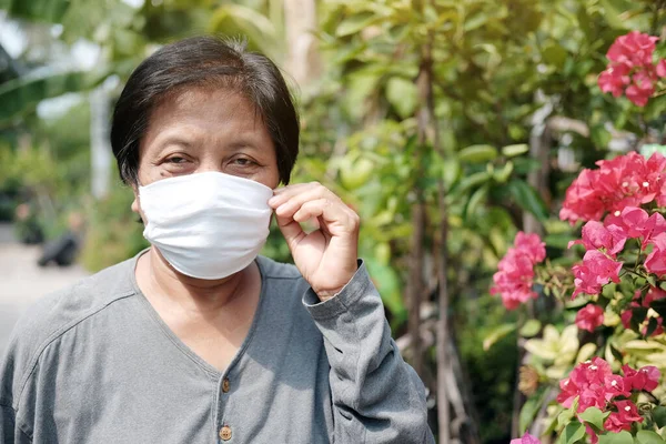 Asian old woman wearing a white cloth mask for prevent the Covid-19 or Corona virus in Thailand and protection for Air Pollution Value Pm 2.5. Health and medicine of elderly in garden concept