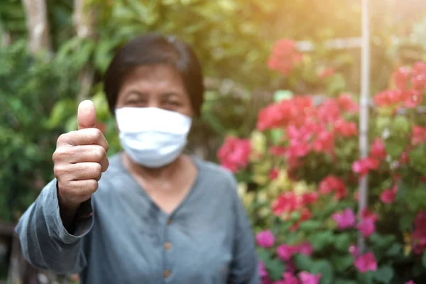 Asian old woman wearing a white cloth mask for prevent the Covid-19 or Corona virus in Thailand and Thumbs up for protection of Air Pollution. Health of elderly concept. Closed up focus on hand.