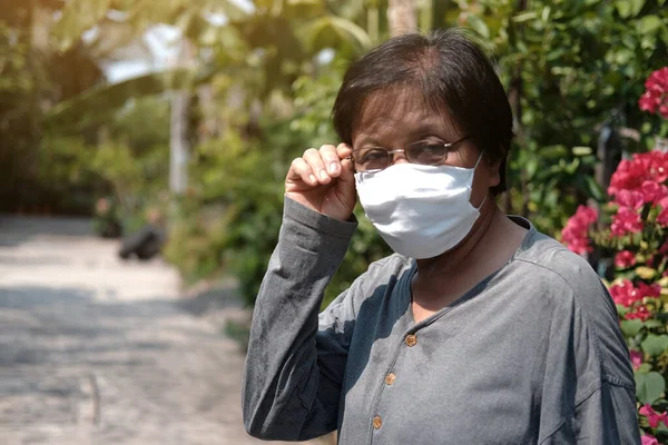 Asian old woman wearing glasses and white cloth mask for prevent the Covid-19 or Corona virus in Thailand and protect for Air Pollution Value Pm 2.5. Health and medicine of elderly in garden concept