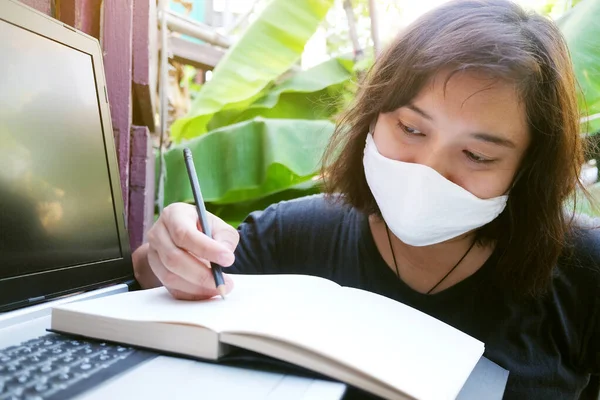 Covid-19 veya Corona virüsünü önlemek için beyaz bez maske takan Asyalı gülümseyen Taylandlı kadın ve evde bilgisayarla çalışmak ve çevrimiçi sistemde çalışmak için yazı yazmak. Hava Kirliliği Değeri Pm 2.5 konsepti.