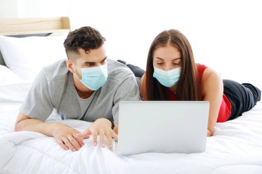Maske takan beyaz bir çift yatak odasındaki sosyal medya teknolojisinde covid-19 virüsünün dizüstü bilgisayarla rahatlamasını engelliyor. Tatilde aile yaşamı için sosyal mesafeler ve sağlık hizmetleri.