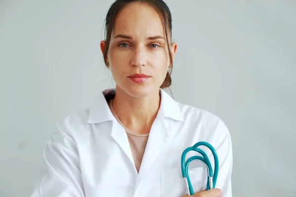 Önlüklü, steteskoplu, beyaz duvar arkasında gülümseyen beyaz saçlı doktor kadın..