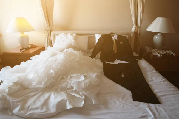 ウェディングドレスとスーツは ホテルの部屋で美しい太陽の光と白いベッドの上に横たわっています バレンタインデーとお祝いのコンセプトへの愛 — ストック写真