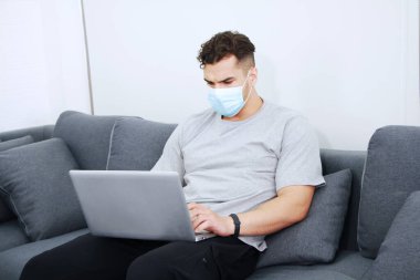 Maske takan genç Kafkas işadamı covid-19 virüsünün rahatlamasını ve aile evindeki oturma odasında sosyal medyada dizüstü bilgisayarla çalışmasını engelliyor. Teknoloji ve iş ev kavramı.