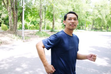 Gülümseyen Asyalı genç adam koşuyor ve bahçede beyaz kulaklıklarla müzik dinliyor..