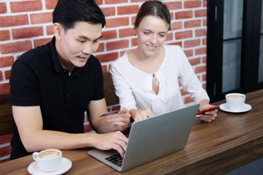 Gülümseyen genç iş adamları Asyalı erkekleri ve Kafkasyalı kadınları sever. Online alışveriş için kredi kartı tutarlar ve sosyal medyada kahve dükkanındaki ahşap masada dizüstü bilgisayarla ödeme yaparlar..