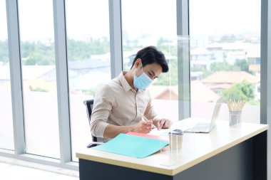 Covid 19 virüsünü önleyen maske takan genç Asyalı işadamı ve ofisteki bilgisayar masasında dizüstü bilgisayarla çalışıyor. Sosyal uzaklık ve yeni normal yaşam tarzı kavramı