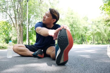 Gülümseyen Asyalı adam çimento zeminde koşup bahçede koştuktan sonra kasları gevşetmek için egzersiz yapıyor ve bacaklarını esnetiyordu. Spor ve sağlıklı kavram