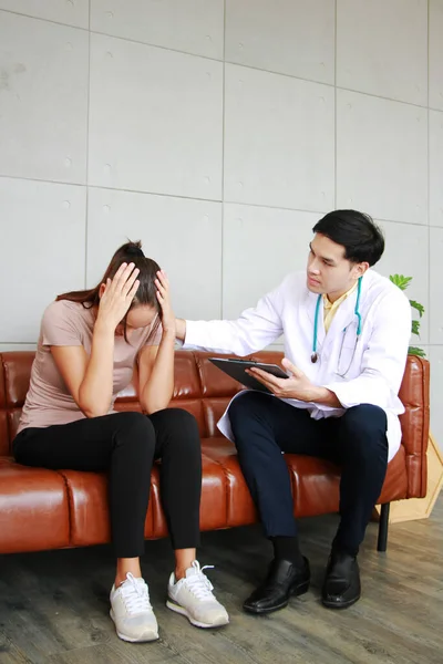 Young asian male psychiatrist is specialist in psychiatry is touching shoulder on Caucasian female Patient is headache on sofa and encouraging to her in Clinical treatment for emotional symptoms.