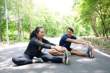 Gülümseyen Asyalı çiftler bahçede beton zeminde koştuktan sonra kaslarını gevşetmek için egzersiz yapıyor ve bacaklarını esnetiyorlar. Sosyal uzaklık ve yeni normal hayat kavramı