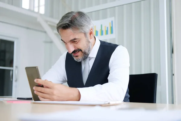 Smiling smart Caucasian senior manager and businessman in black suit relaxing in social media online and talking, calling with businesspeople on smartphone at workspace in office