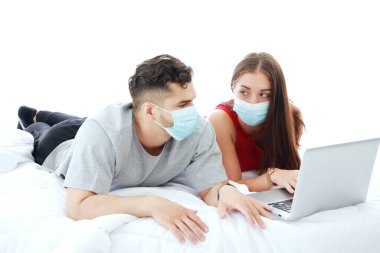 Maske takan beyaz bir çift covid-19 virüsünün yatak odasındaki sosyal medya teknolojisinde dizüstü bilgisayarla yatmasını engelliyor. Tatilde evli aile hayatı için sosyal mesafeler ve sağlık hizmetleri.