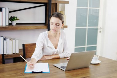 Başarılı Kafkasyalı genç iş kadını internette dizüstü bilgisayarla araştırma yapıyor ve ofisteki işyerindeki kağıt üzerinde bilgi ve bilgiler yazıyor. Eğitim ve Ticaret Teknolojisi.