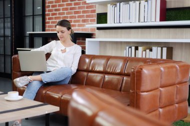 Kahverengi kanepede rahatlayan ve oturma odasında dizüstü bilgisayarla iletişim kuran gülümseyen beyaz bir aile. İş kadınlarının teknoloji ve yaşam tarzı tatil konsepti üzerine