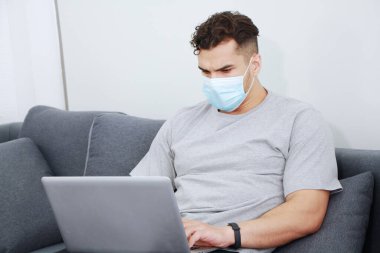 Maske takan genç Kafkas işadamı covid-19 virüsünün rahatlamasını ve aile evindeki oturma odasında sosyal medyada dizüstü bilgisayarla çalışmasını engelliyor. Teknoloji ve iş ev kavramı.