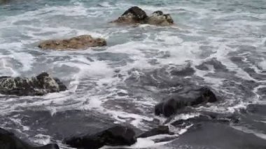 Deniz dalgaları kayalardan oluşan karanlık kayalara çarpar. Köpüklü sörf.