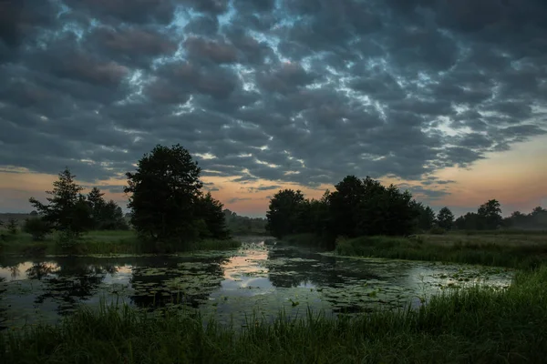 Salida del sol nebulosa sobre un prado y un río con nubes oscuras en el cielo . — Foto de Stock