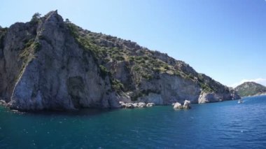 Kayalık bir deniz kıyısı ve beyaz bir tekne dalgalarda yüzüyor..