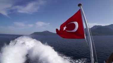 Türkiye bayrağı, mavi denizin, dağların ve geminin motorunun köpük izlerine karşı zevk teknesinin kıç tarafında dalgalanır..