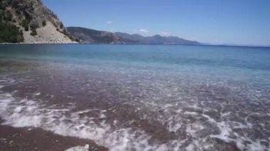 Çakıl taşlı sahilde şeffaf deniz dalgaları, ufukta dağlar.