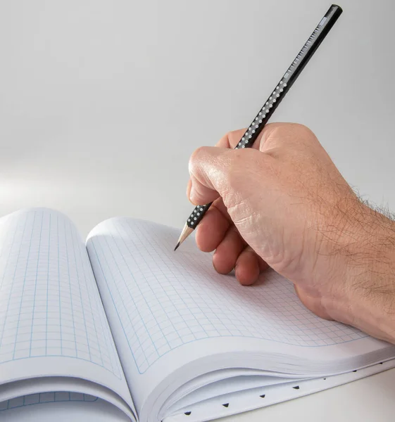 Male hand with a pencil writes in a checkered notebook sheet. Selective focus. Blurred background.