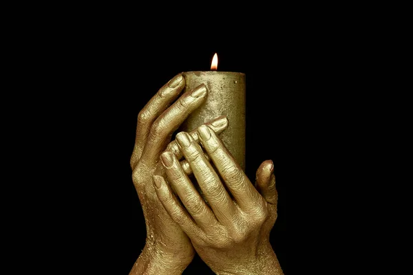 Lit gold candle in golden hands on black background. Occult ritual symbol of wealth and well-being.