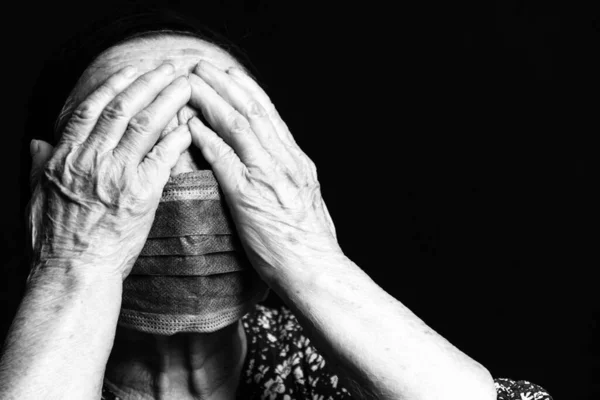 Black and white portrait of an elderly woman in medical mask covering her eyes with her hands on a black background. Old people in risk zone because of virus epidemy, COVID-2019 pandemic