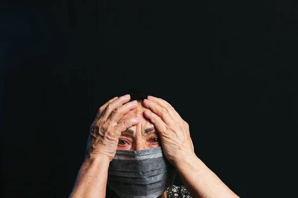 Siyah arka planda iki eli tıbbi maske takmış yaşlı bir kadının portresi. Virüs salgını, COVID-2019 salgını yüzünden risk altındaki yaşlılar
