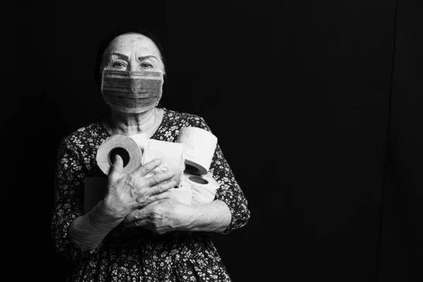 Black and white portrait of an elderly woman in medical mask holding rolls of toilet paper on a black background. COVID-2019 hoarding fever
