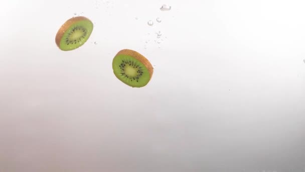 Slices of kiwi fruit slowly fall into the clear water on a white background — Stock Video