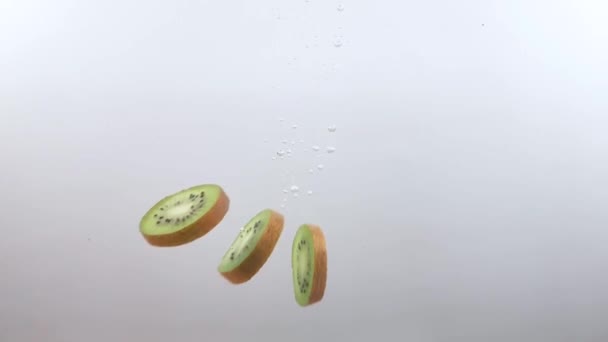 Slices of kiwi fruit slowly fall into the clear water on a white background — Stock Video