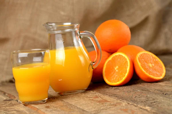 Стекло с апельсиновым соком, кувшин со свежим соком и куча апельсинов на заднем плане на деревянном столе — стоковое фото
