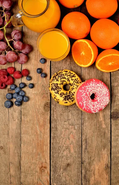Donuts, suco de laranja, mirtilos, framboesas, maçãs, laranjas - café da manhã doce na mesa de madeira - visão de cima, foto vertical — Fotografia de Stock