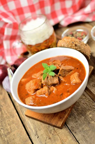 Close-up of pork goulash with pieces of meat in a bowl, garlic, pepper, onion, glass with cold beer and red checkered tablecloth in the background - vertical photo — Stock Photo, Image