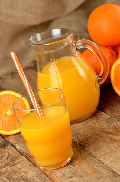 Vaso con zumo de naranja y paja, jarra con zumo fresco y pila de naranjas en el fondo sobre mesa de madera — Foto de Stock