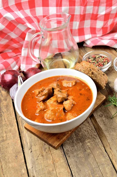 Gulasch di maiale con pezzi di carne in una ciotola, aglio, pepe, cipolla, brocca con olio e tovaglia a quadretti rossi sullo sfondo - foto verticale — Foto Stock