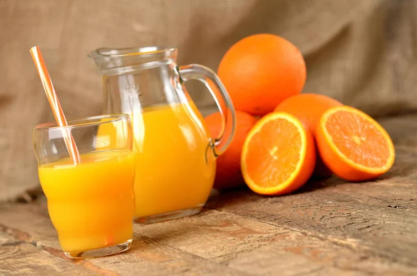Стекло с апельсиновым соком и соломой, кувшин со свежим соком и куча апельсинов на заднем плане на деревянном столе — стоковое фото