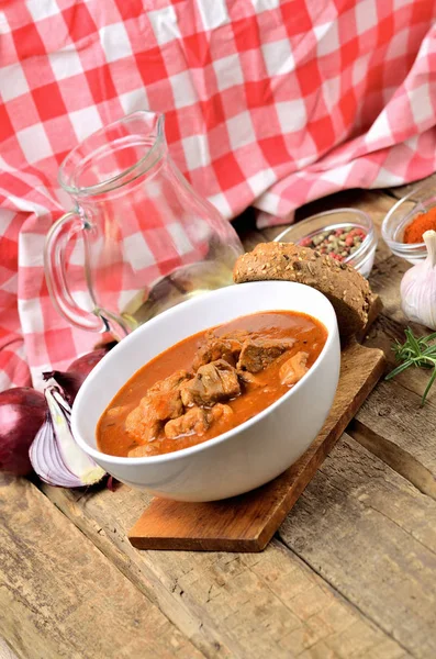 Pork goulash with pieces of meat in a bowl, garlic, pepper, onion, jug with oil and red checkered tablecloth in the background - vertical photo — Stock Photo, Image