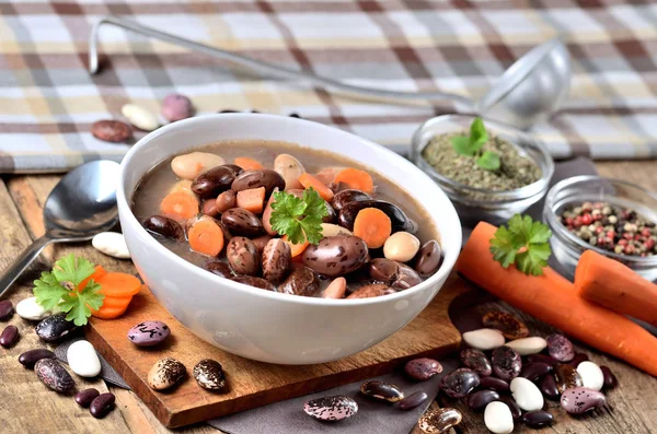 Bowl of bean soup with large beans on cutting board, carrots, parsley, marjoram, spoon, towel and ladle in background