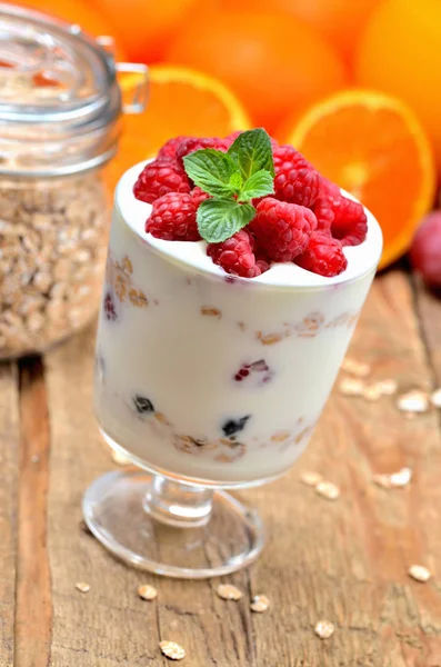 Griechischer Joghurt mit Haferflocken und frischen Himbeeren im Glas und Minzblättern, komplettes Glas Haferflocken und Orangen im Hintergrund — Stockfoto