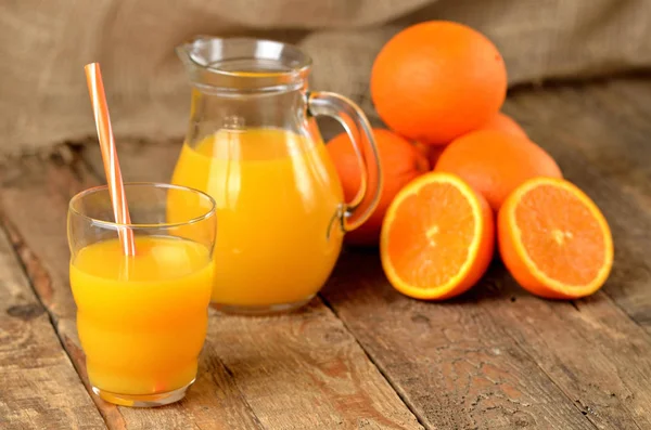 Ποτήρι με χυμό πορτοκάλι και άχυρο, κανάτα με φρέσκο χυμό και σωρό πορτοκάλια στο παρασκήνιο στο ξύλινο τραπέζι — Φωτογραφία Αρχείου