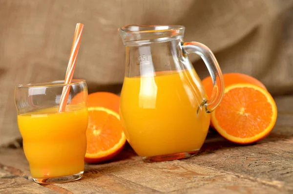 Vaso lleno de zumo de naranja, jarra con zumo fresco, paja y naranjas frescas en el fondo sobre mesa de madera — Foto de Stock