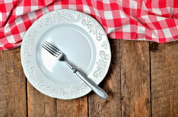 Beyaz dekoratif seramik tabak ve eski vintage ahşap masa arka plan üzerinde üst tarafında kırmızı kareli masa örtüsü çatal - yukarıdan görüntüleyin — Stok fotoğraf