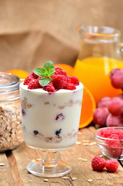 Griechischer Joghurt, frische Himbeeren im Glas und Minzblätter, Orangen, Trauben und ein volles Glas Haferflocken im Hintergrund - gesundes Frühstückskonzept — Stockfoto