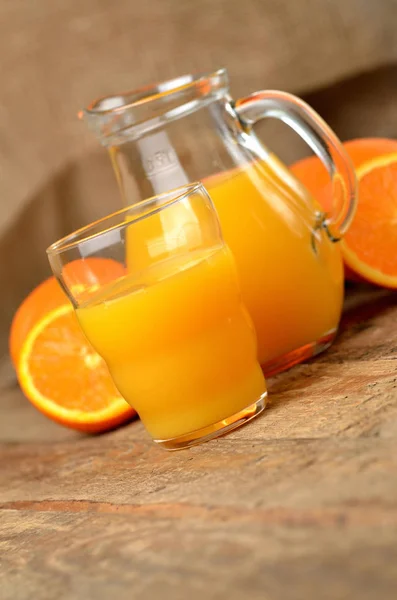 Стекло, полное апельсинового сока, графин со свежим соком и свежими апельсинами на заднем плане на деревянном столе - вертикальное фото — стоковое фото